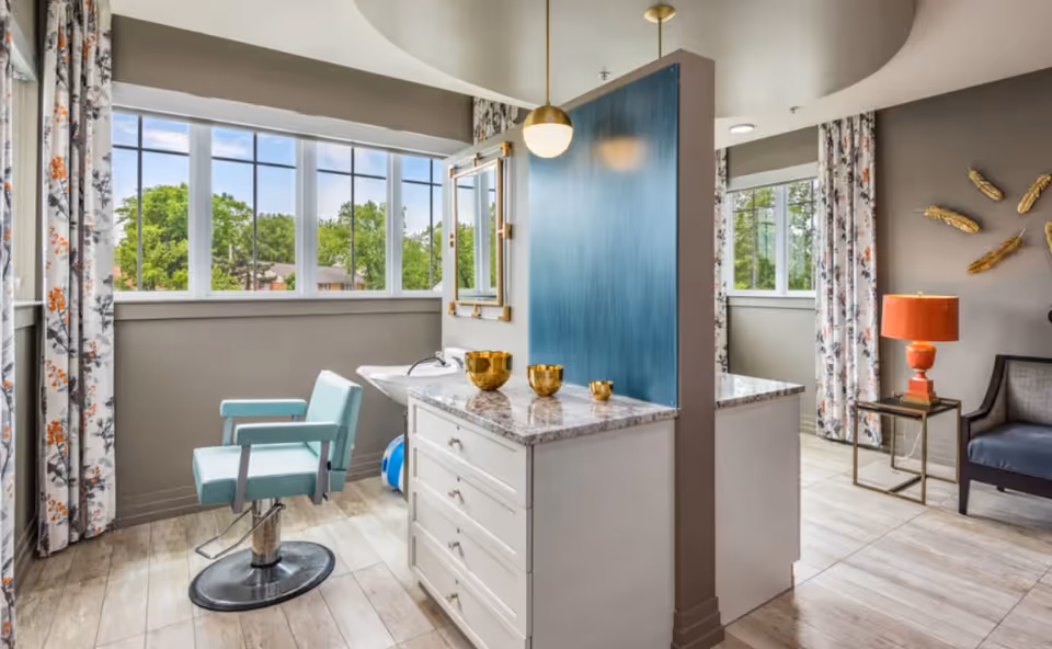 Bright interior salon-style grooming station with a turquoise barber chair, marble-topped vanity, large windows, and an adjacent seating area with a lamp.