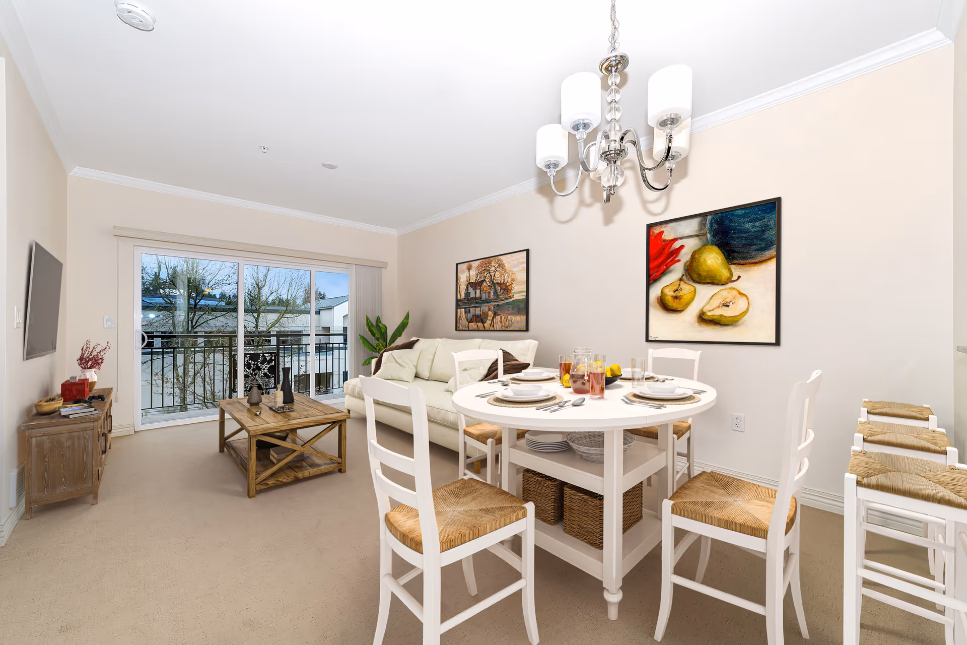 A bright living and dining area with a white round dining table set for four, white chairs with woven seats, a beige sofa, a wooden coffee table, and a TV mounted on the wall. Large sliding glass doors open to a balcony with a view of trees and neighboring buildings. Two framed paintings hang on the walls, and a chandelier with five lights hangs above the dining table.