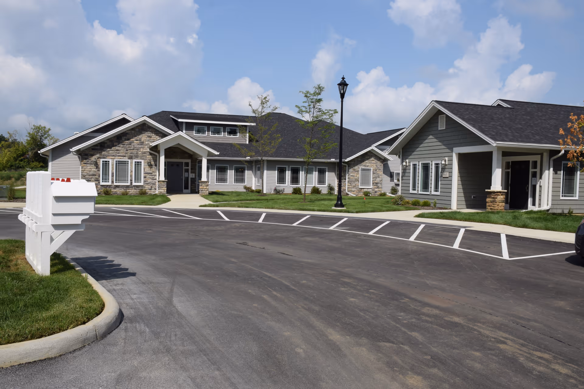 Exterior view of a senior living facility building with gray siding and stone accents, a paved driveway with parking spaces, a white mailbox cluster on the left, and a lamppost in the center under a partly cloudy sky.