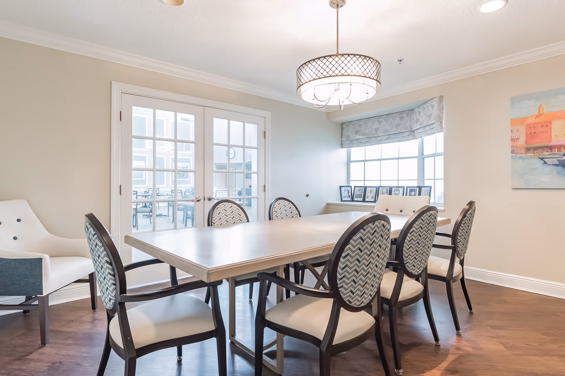 A bright dining room with a rectangular table surrounded by six upholstered chairs with patterned backs and cream seats. There is a white armchair with dark legs in the corner. The room has wooden flooring, beige walls, and a large window with a patterned valance. A modern chandelier hangs above the table, and a painting is visible on the right wall. French doors with glass panes lead to another room with additional tables and chairs.