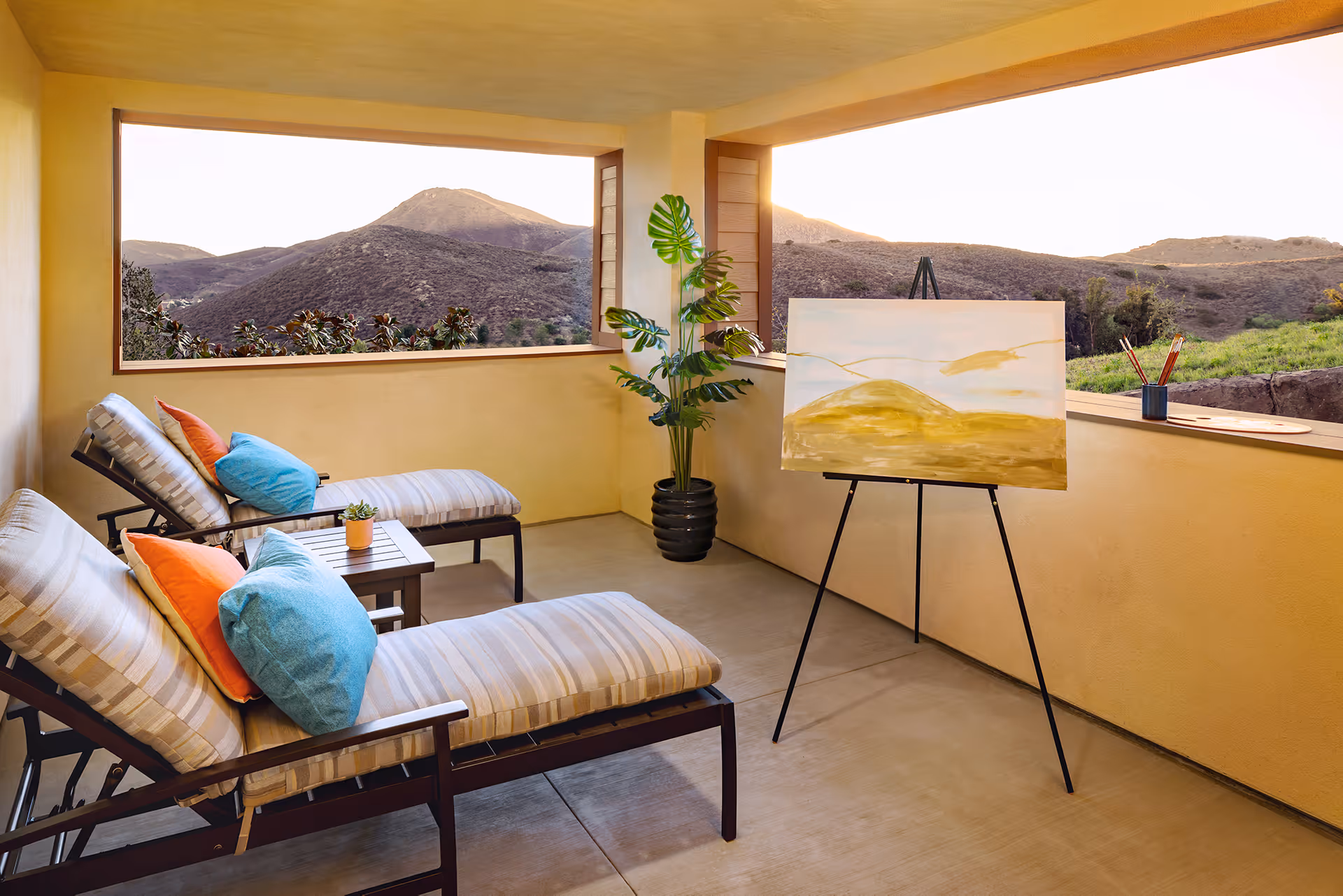 Covered outdoor patio area with two cushioned lounge chairs featuring blue and orange pillows, a small side table with a potted plant, a large potted plant in the corner, and an easel holding a painting of a landscape. The patio overlooks a scenic view of hills and mountains in the background.