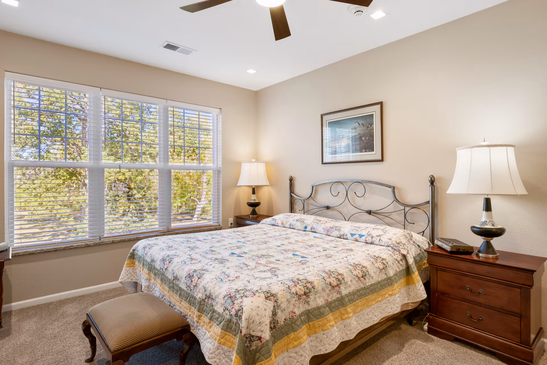 Bright furnished bedroom with a large window, a floral-quilted bed, bedside tables and lamps.