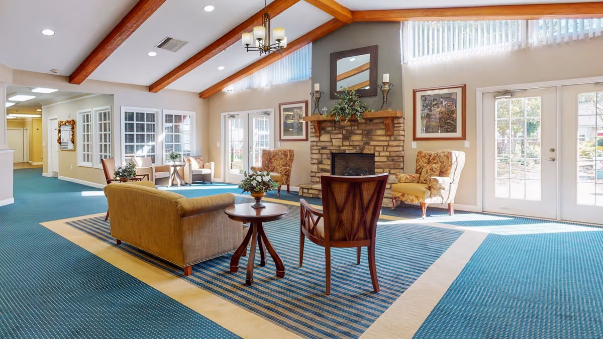 Spacious senior living common room with seating around a stone fireplace, high beamed ceiling, and large windows and glass doors letting in sunlight.