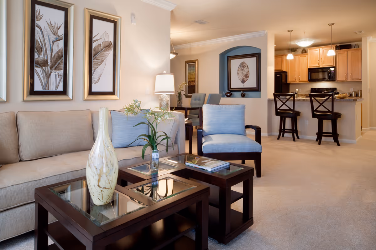 A cozy living room area with a beige sofa, a light blue armchair, and two dark wooden glass-top coffee tables. The tables have decorative items including a tall vase and a small plant. The room features framed botanical artwork on the walls, a table lamp, and an open view into a kitchen with wooden cabinets and two bar stools at a counter. The carpet is light beige, and the overall decor is warm and inviting.