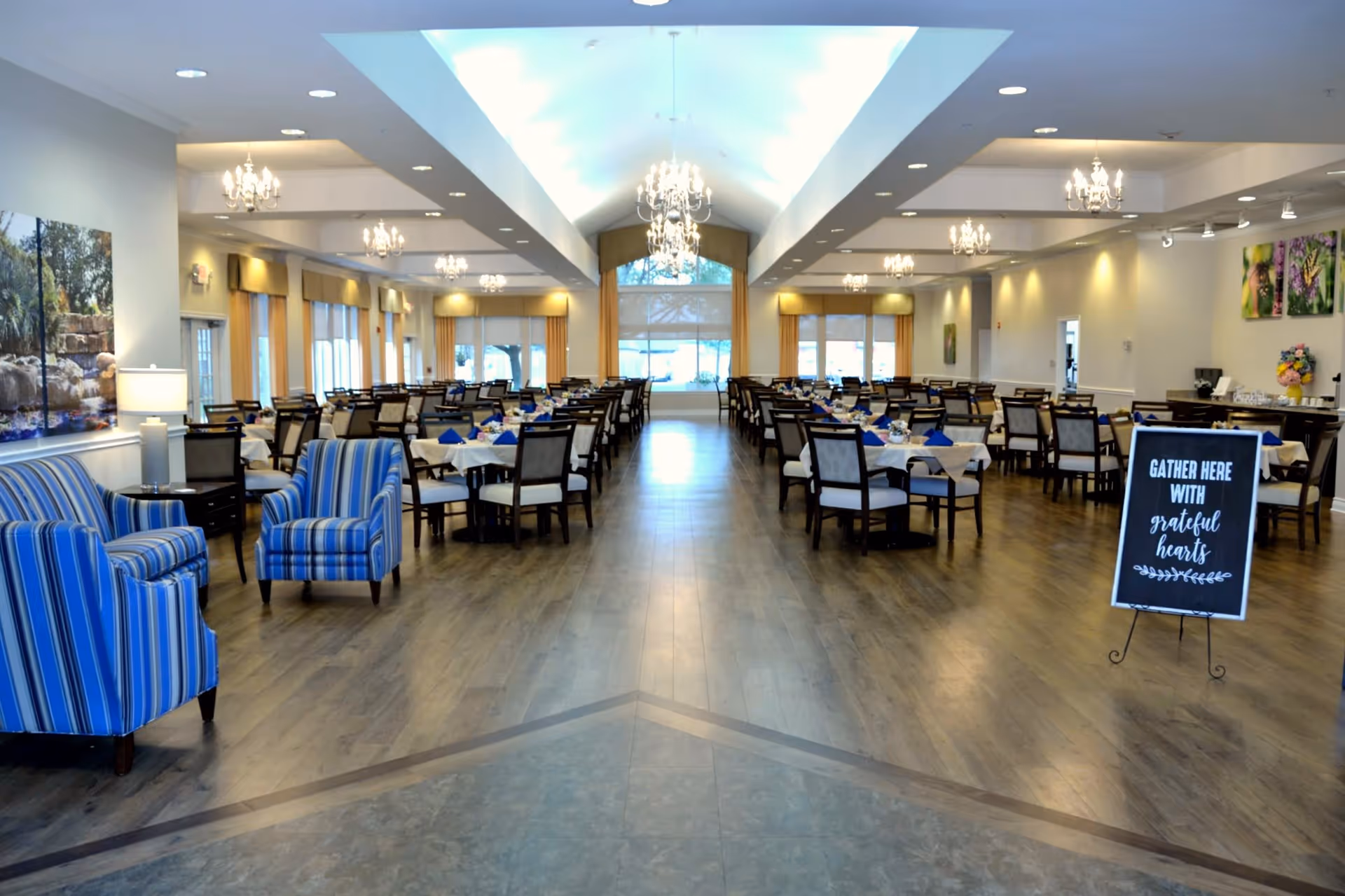 Spacious dining room with multiple tables and chairs neatly arranged, blue-striped armchairs on the left, chandeliers hanging from the ceiling, large windows with curtains, and a sign that reads 'Gather here with grateful hearts'.