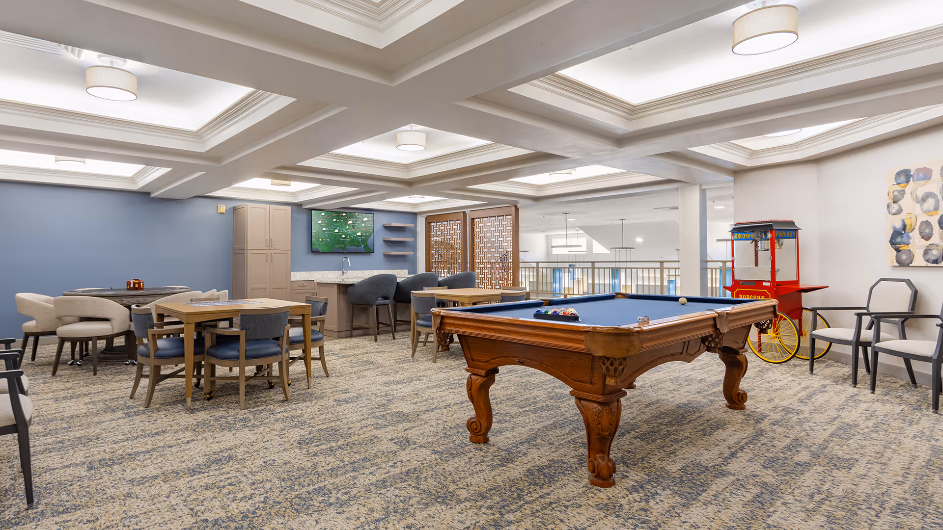 Spacious senior living common room with a pool table, multiple seating areas, and a popcorn cart under coffered ceilings.
