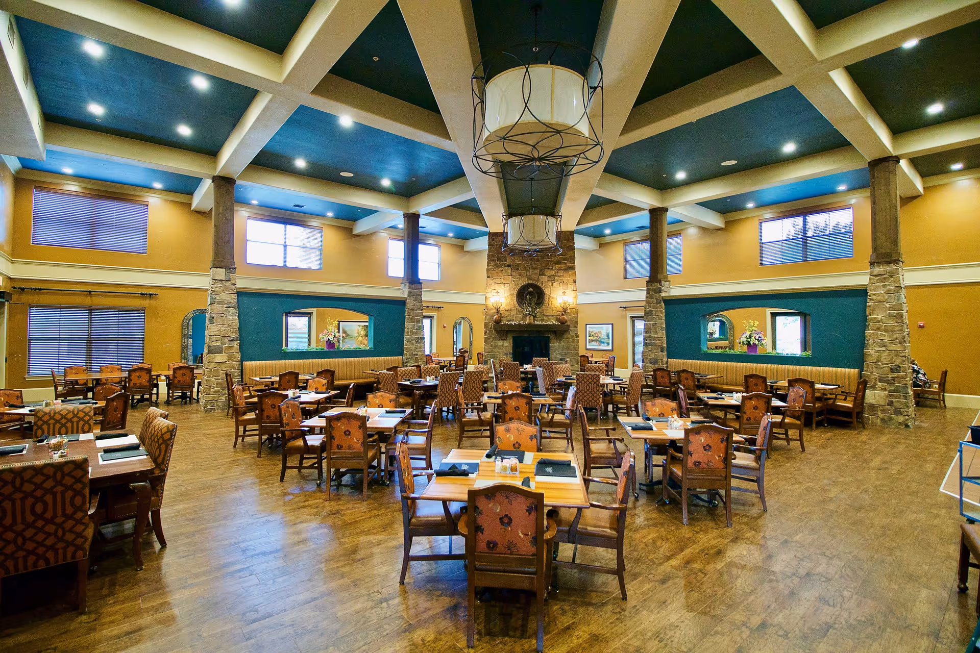 A spacious dining room with multiple wooden tables and chairs arranged neatly. The room features a high ceiling with exposed beams painted blue and beige, stone pillars, large windows allowing natural light, and a central stone fireplace. The walls are painted in warm tones with teal accent sections, and there are decorative light fixtures hanging from the ceiling.