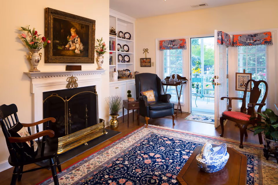 A cozy living room with a fireplace, two armchairs, a wooden rocking chair, and a coffee table with a blue and white ceramic decoration. The room has a large patterned rug, built-in shelves with decorative plates, and a painting above the fireplace. French doors open to a patio with outdoor furniture and greenery visible outside.