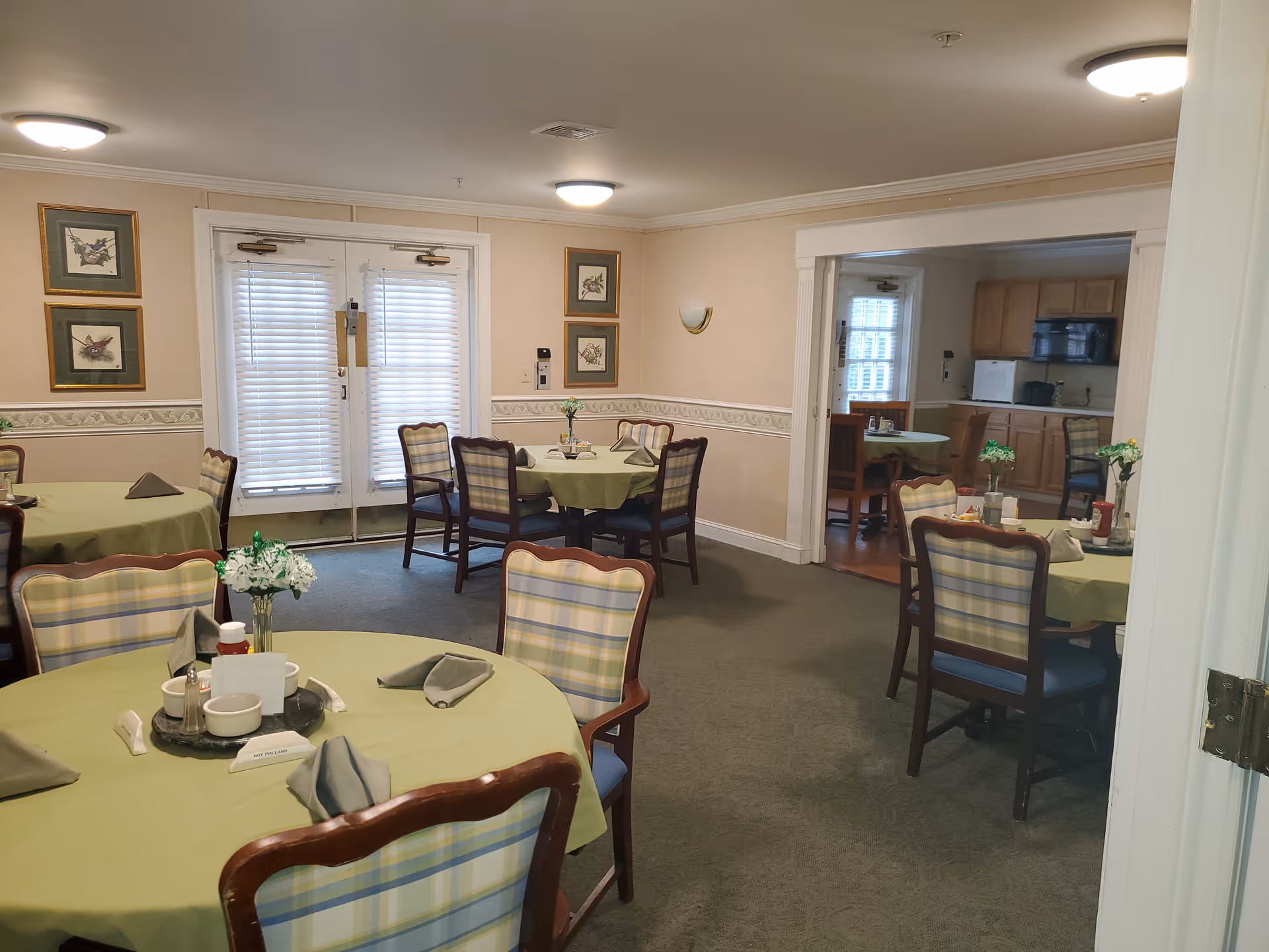 Interior view of a dining room in a senior living facility with round tables covered in green tablecloths, each set with napkins, condiments, and small flower vases. The room has plaid upholstered chairs, framed artwork on the walls, and a doorway leading to a kitchen area with wooden cabinets and appliances.