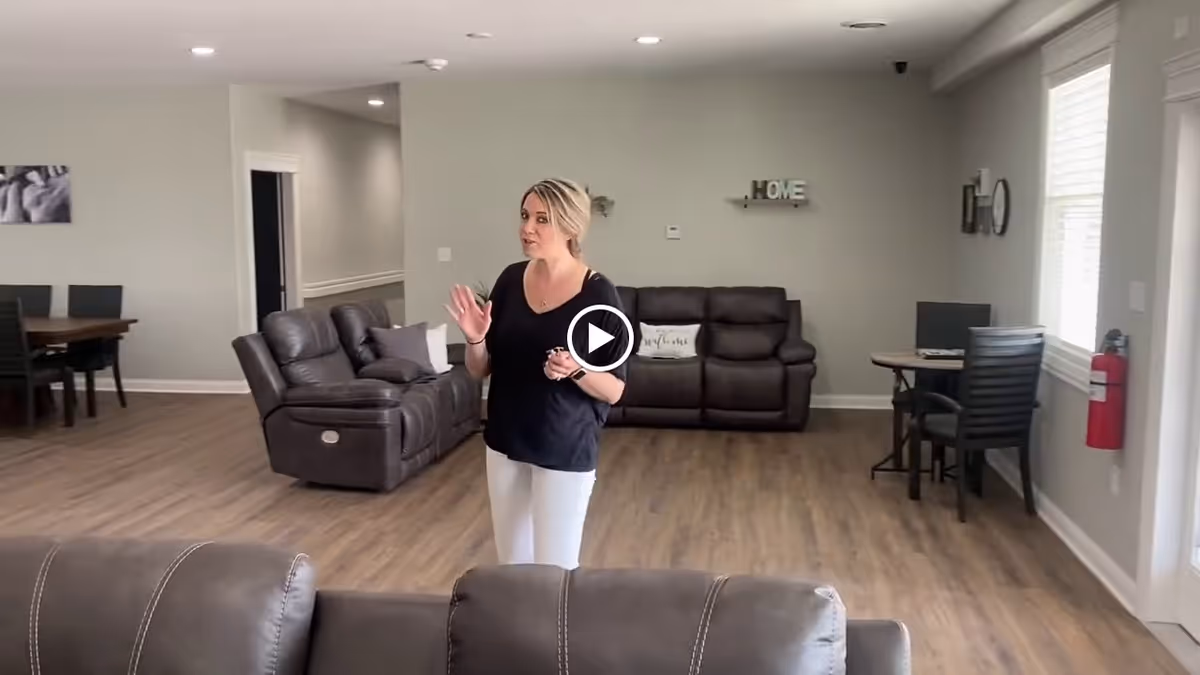 A woman standing in a spacious living room area with brown leather sofas, wooden flooring, and light-colored walls. There is a dining table with chairs in the background to the left, a small desk with a chair and computer to the right near a window, and a fire extinguisher mounted on the wall. The room has a calm and welcoming atmosphere with a decorative 'HOME' sign on the wall behind the woman.