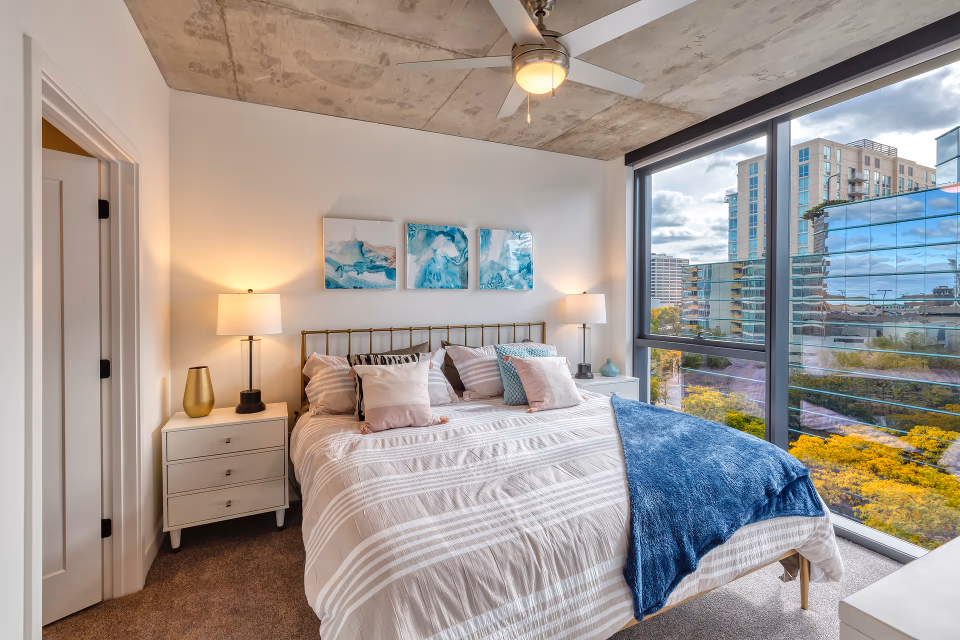 Modern bedroom with a bed, bedside tables and lamps, artwork above the headboard, and a large floor-to-ceiling window showing a city view.