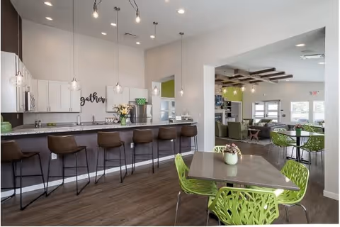 Modern communal kitchen and dining area with a long counter featuring six brown bar stools, white cabinets, pendant lights, and a sign that says 'gather'. In the foreground, there is a table with four green chairs and a small plant centerpiece. In the background, a living area with green chairs and sofas is visible through an open doorway.