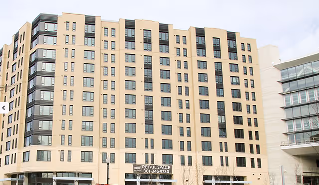 Exterior view of a multi-story beige building with many windows, featuring a sign for retail space leasing with a phone number. The building has a modern design with a corner section of dark-framed windows and is adjacent to another building with glass balconies.