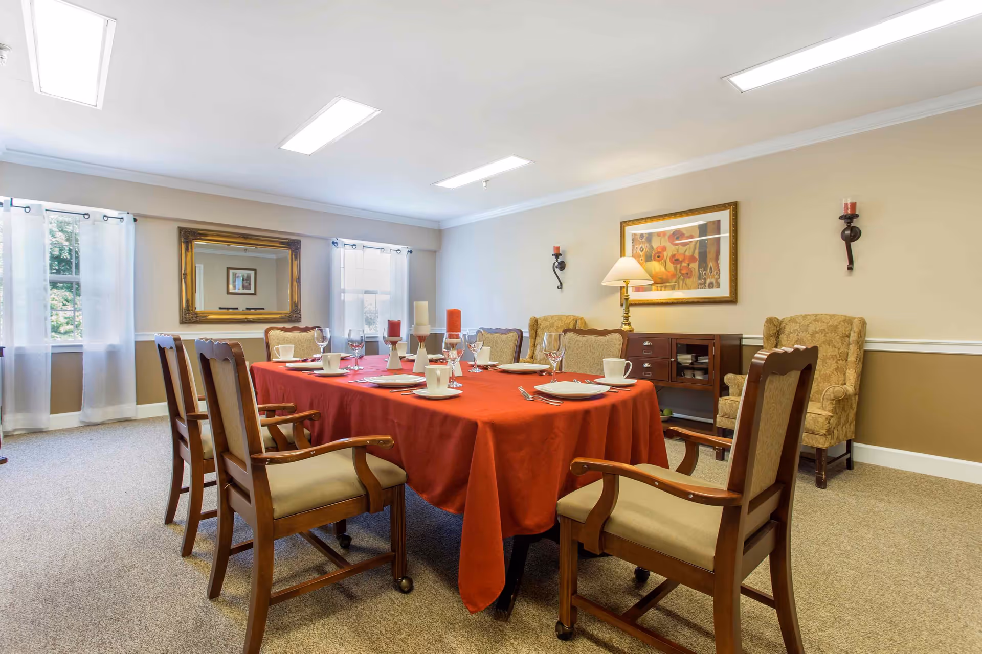 A dining room with a rectangular table covered with a red tablecloth, set with plates, cups, glasses, and candles. The room has beige walls, carpeted floor, two windows with sheer white curtains, a large mirror on one wall, two upholstered armchairs, a wooden sideboard with a lamp, and a framed painting above it.