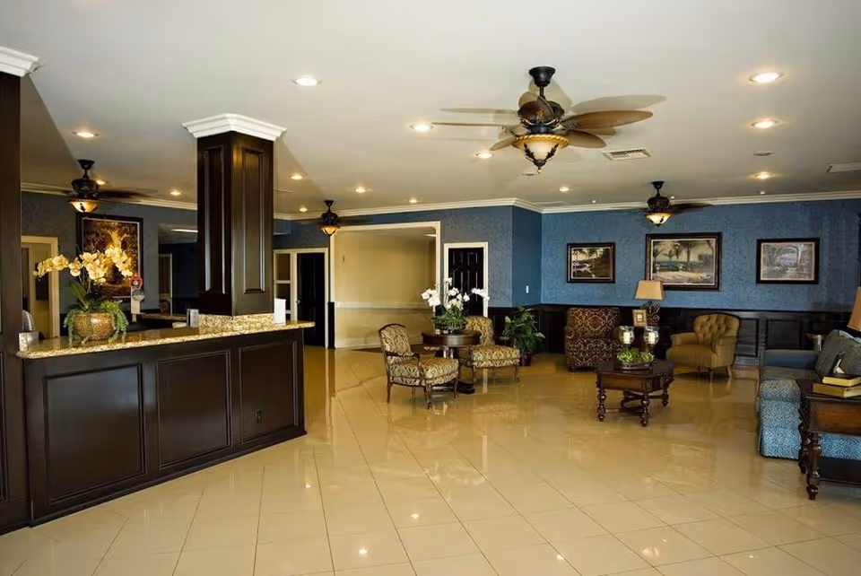 A spacious and well-lit interior common area with polished tile floors, dark wood furniture, and blue walls. The room features a reception desk with a granite countertop, several upholstered chairs and sofas arranged around wooden coffee tables, ceiling fans with lights, framed artwork on the walls, and decorative plants.