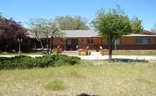 Single-story brick building with a patio area in front, featuring outdoor seating and surrounded by trees and grass under a clear blue sky.