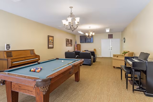 A spacious community recreation room with a pool table, pianos, couches and a wall-mounted TV.
