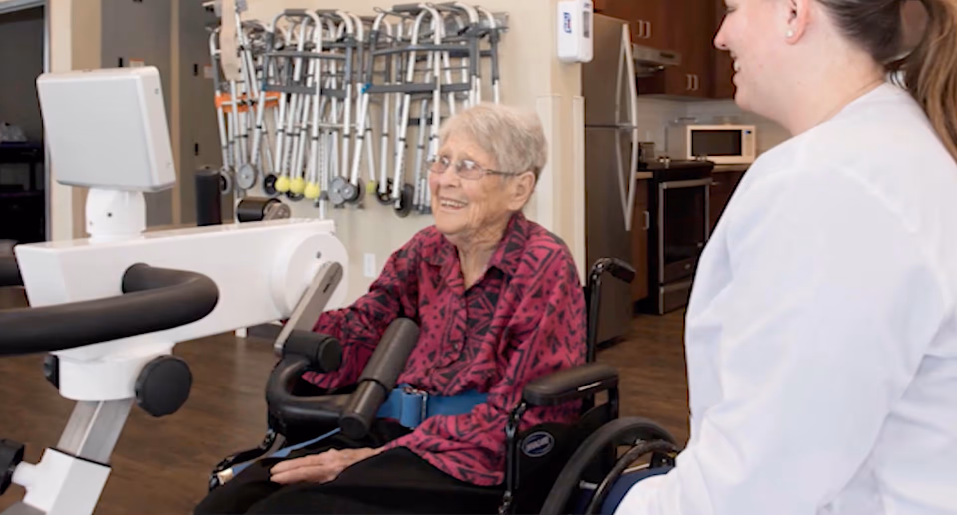 An elderly woman in a wheelchair using an exercise machine with the assistance of a healthcare worker in a room with walkers hanging on the wall and a kitchen area in the background.