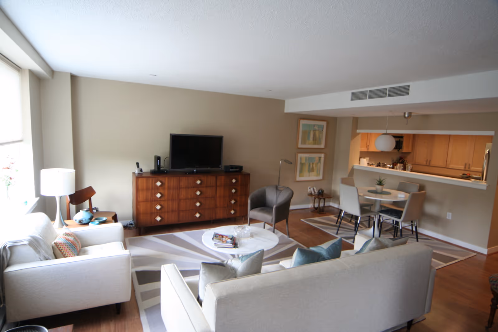 Bright open living room with sofas, a TV on a wooden dresser, round coffee table, and an adjoining dining area with a kitchen pass-through.