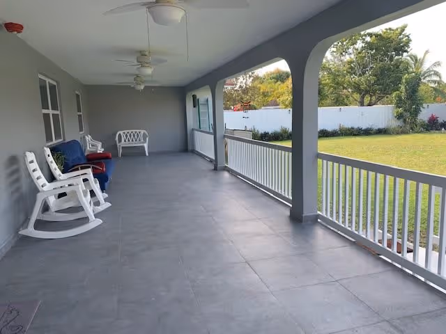 A spacious covered porch with gray tiled flooring and white railings overlooking a green lawn and garden. The porch has several white rocking chairs and a bench, ceiling fans, and a gray wall with windows and a door.