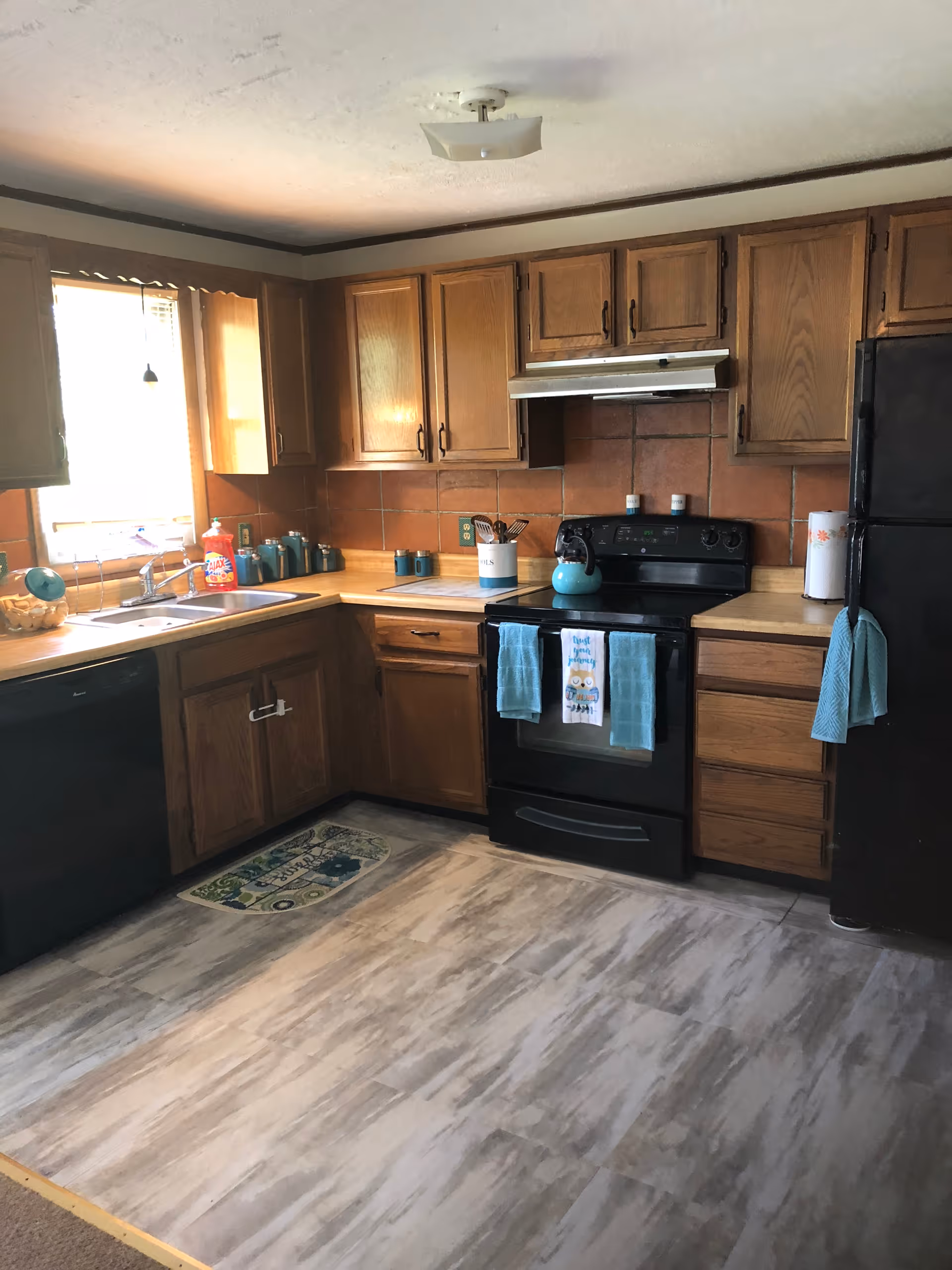 A kitchen with wooden cabinets, a black stove with a teal kettle on it, a black refrigerator, and a dishwasher. The countertops are light-colored, and there are teal kitchen towels hanging on the stove and refrigerator. A window above the sink lets in natural light, and there are various kitchen items like a dish soap bottle and canisters on the counters. The floor has a light wood-like pattern.