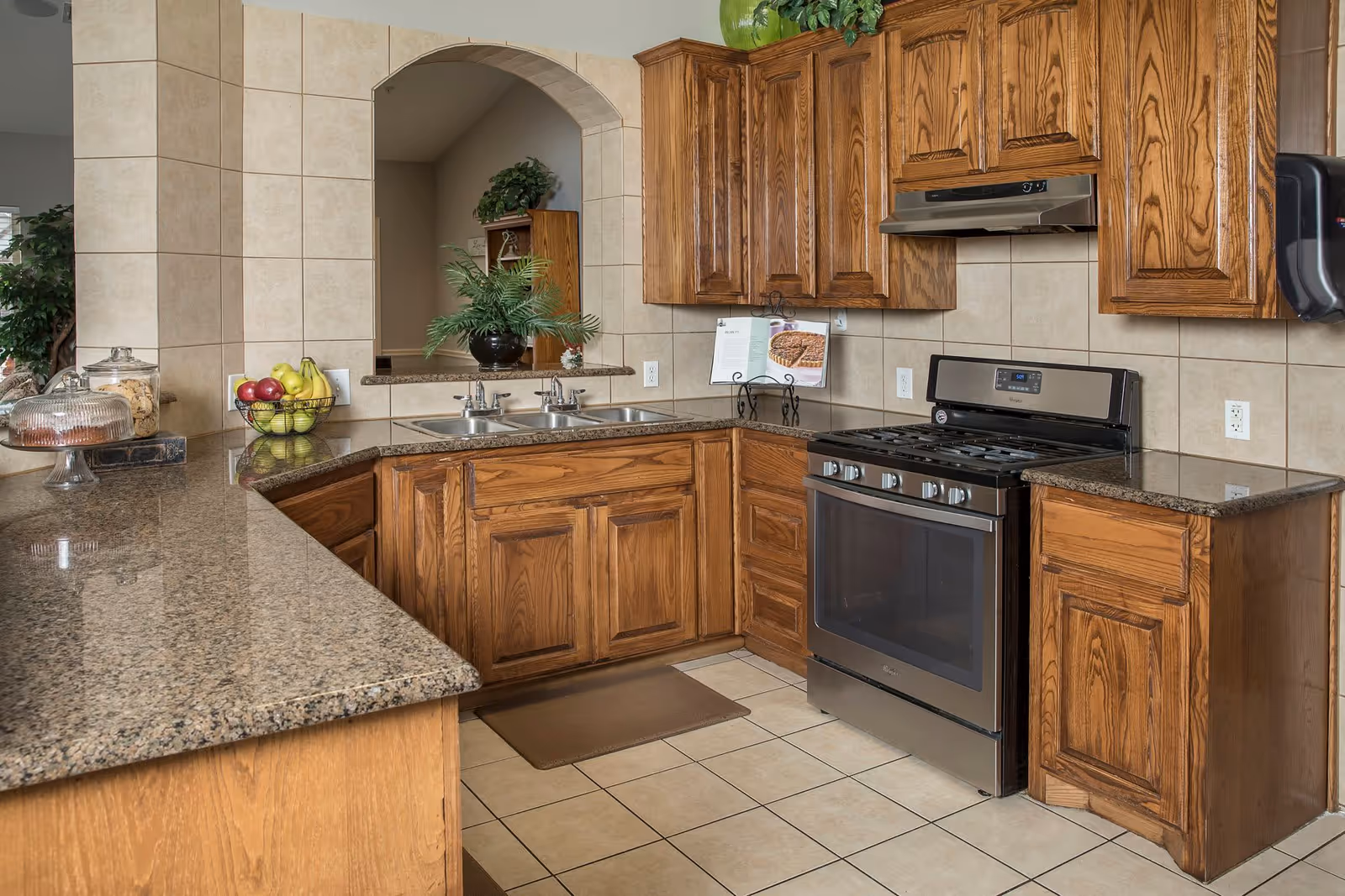 A kitchen with wooden cabinets, a granite countertop, a stainless steel gas stove, and a double sink. There is a bowl of fruit and a glass cake stand on the counter, with a plant visible through an arched opening in the tiled wall.
