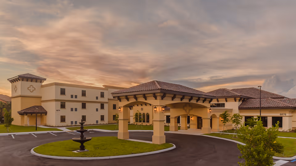 Exterior view of Discovery Commons Cypress Point facility at sunset, showing a beige building with a tiled roof, a covered entrance with columns, a circular driveway with a fountain in the center, and a well-maintained lawn.