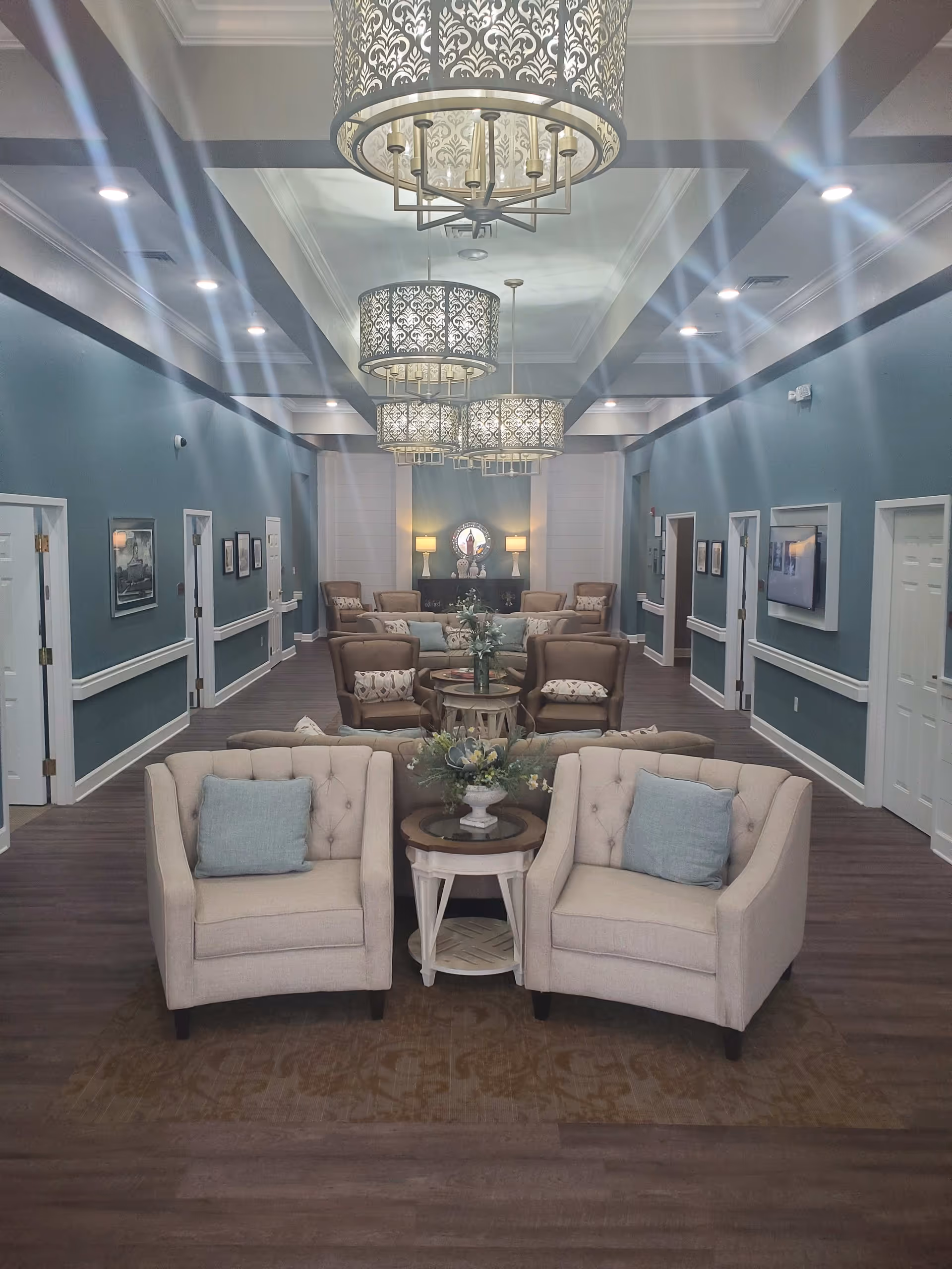 A spacious and well-lit lounge area with multiple beige armchairs and sofas arranged around small tables with floral centerpieces. The walls are painted teal with white trim, and several decorative light fixtures hang from the ceiling. The room has a warm and inviting atmosphere with framed pictures on the walls and a mirror with lamps on a console table at the far end.