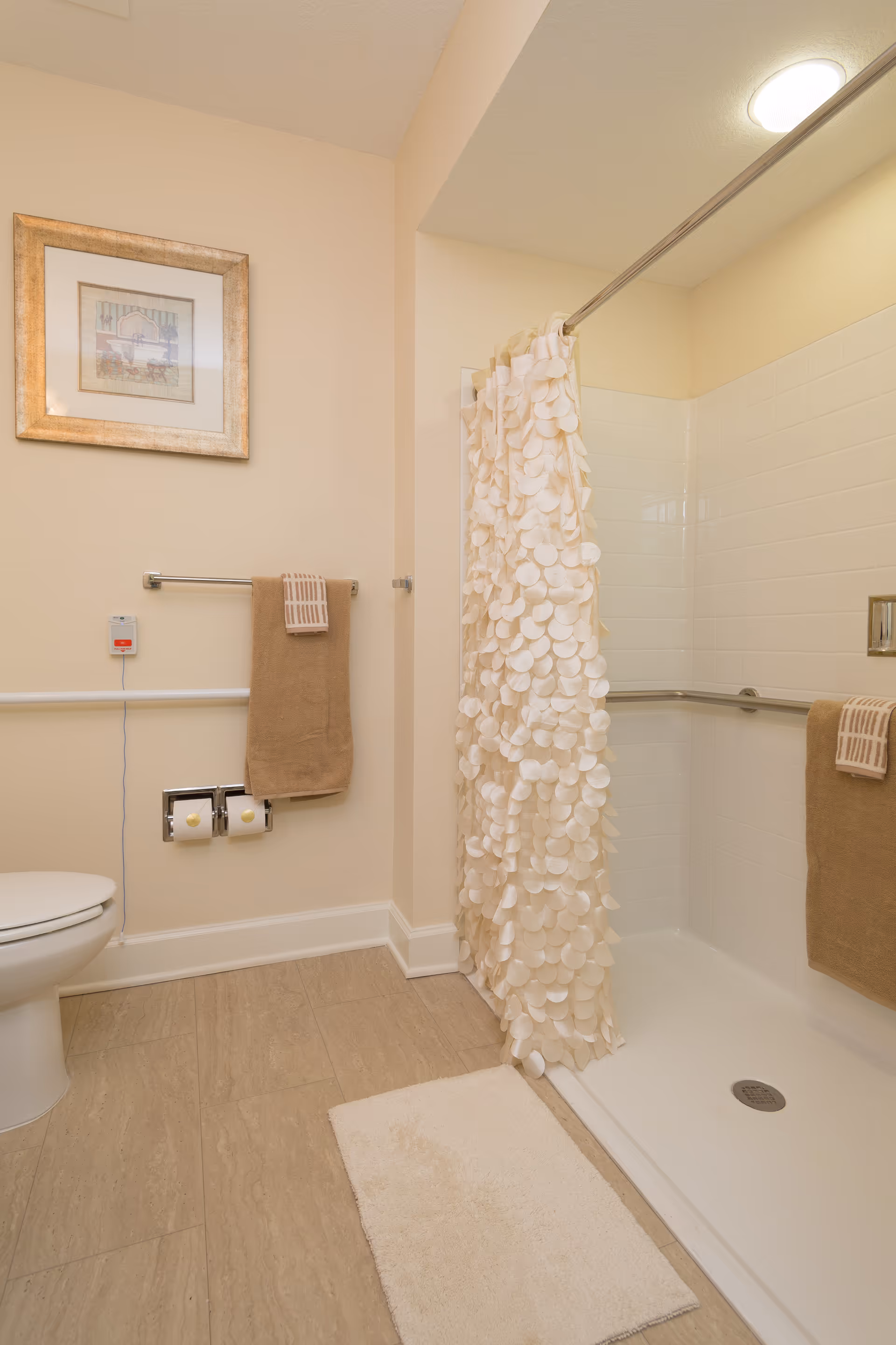 A clean and well-lit bathroom featuring a white toilet, a shower with a white textured curtain, beige walls, and beige floor tiles. There are two beige towels hanging on metal bars, a white bath mat on the floor, a framed picture on the wall, and a grab bar installed inside the shower for safety.