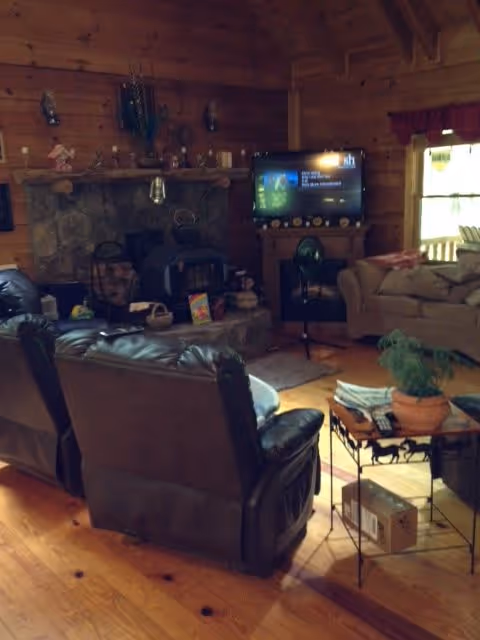 Cozy living room with wooden walls and ceiling, featuring a stone fireplace with a wood stove insert. Two dark leather recliners face a television on a wooden stand. A beige couch is positioned near a window with red curtains. A small table with a potted plant, magazines, and a cardboard box is in the foreground on a wooden floor.