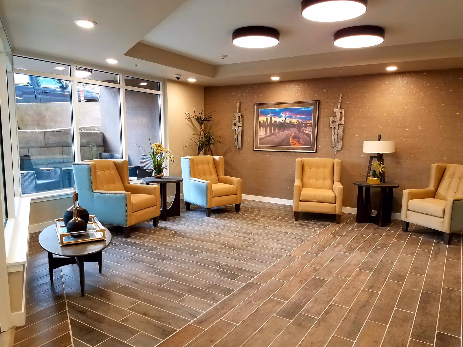 A modern seating area with several beige armchairs, round side tables, decorative plants and wall art beside large windows.