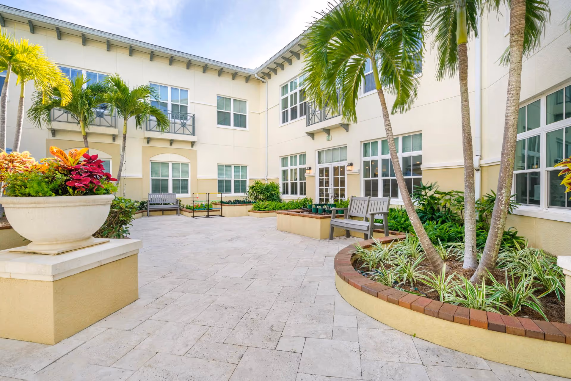 Sunlit courtyard with palm trees, benches, potted plants, and surrounding two-story building windows.