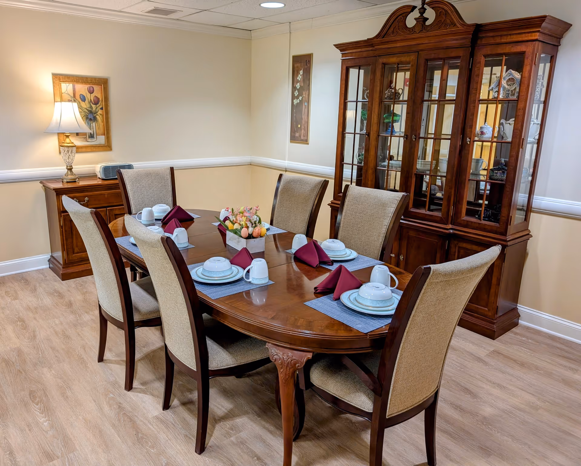 A formal dining room with a wooden dining table set for six people. The table has beige cushioned chairs, white plates, cups, and maroon folded napkins on blue placemats. A floral centerpiece decorates the middle of the table. In the background, there is a wooden china cabinet with glass doors displaying decorative items, and a wooden side table with a lamp and framed artwork on the wall.