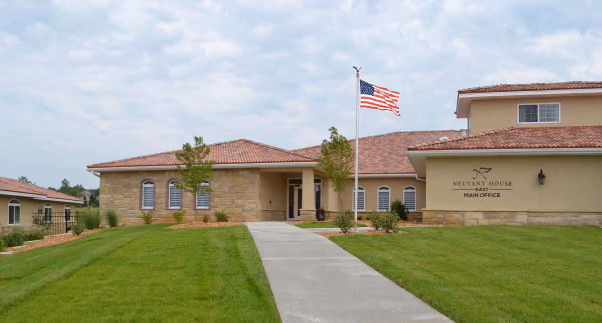 Exterior view of Neuvant House of Lawrence main office building with a stone and stucco facade, a tiled roof, an American flag on a flagpole, and a concrete walkway leading to the entrance surrounded by green grass and small trees.