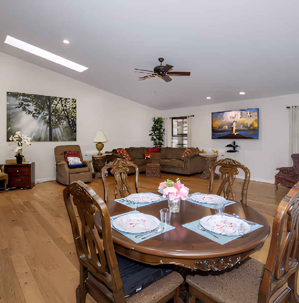 A spacious living and dining area with a wooden dining table set for four, featuring plates, napkins, and a vase with pink flowers. The room has hardwood floors, a ceiling fan, a large brown sectional sofa, an armchair, side tables with lamps, and artwork on the walls. There is a window with curtains and a tall green plant in the corner.