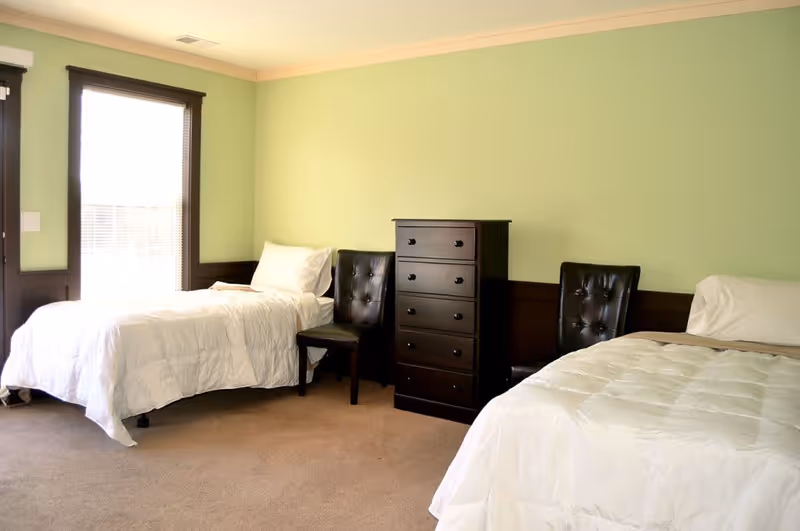 A bright bedroom with two single beds, each with white bedding and pillows. Between the beds is a dark wooden chest of drawers and two dark brown tufted chairs. The walls are painted light green with dark wood trim, and there is a large window with blinds letting in natural light.
