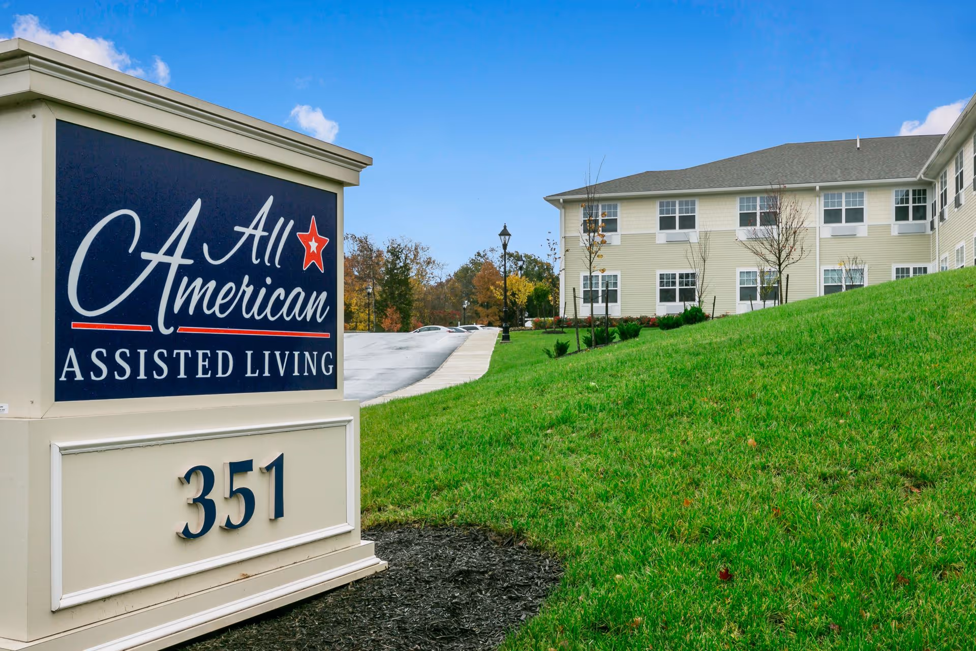 Outdoor view of All American Assisted Living at Wareham showing a large sign with the facility name and address number 351, with a two-story building and green lawn in the background under a blue sky with some clouds.