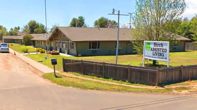 Exterior view of Birch Assisted Living facility showing a single-story building with a brown roof and green walls, surrounded by a wooden fence and a grassy lawn. A sign in front displays the facility's name and contact number. Trees and a clear sky are visible in the background.