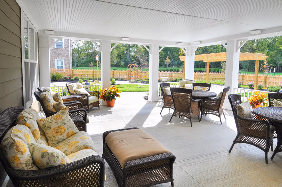 Covered outdoor patio area with wicker furniture including cushioned chairs, sofas with floral pillows, and round tables with chairs. The patio overlooks a fenced garden with green grass, plants, and trees in the background under a clear sky.