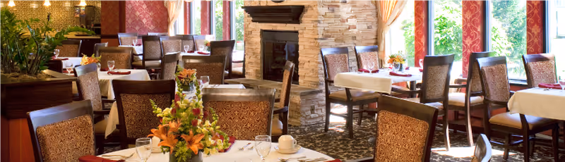 Bright, elegant dining room with multiple set tables, upholstered chairs, floral centerpieces, and a stone fireplace by large windows.
