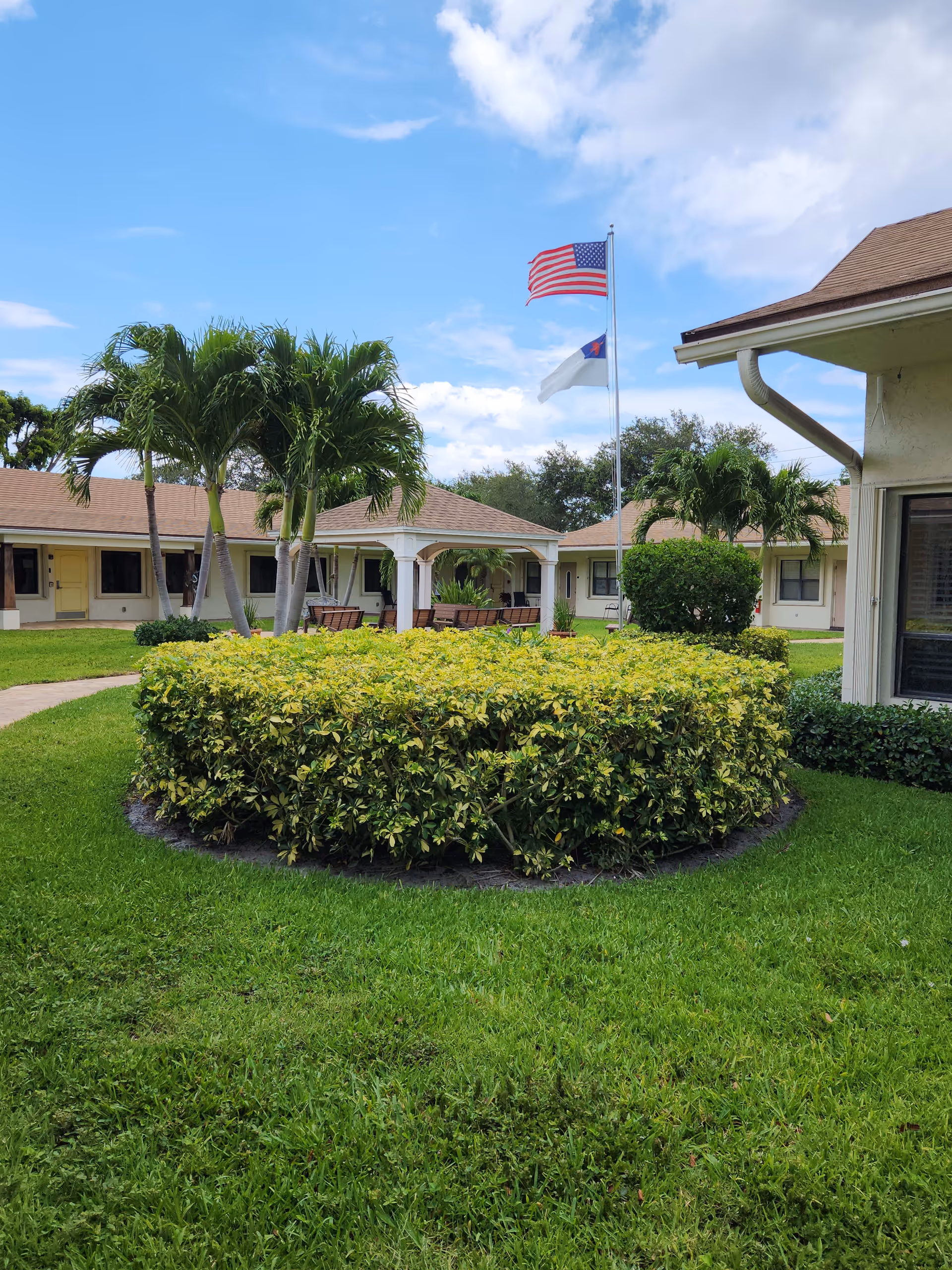 Outdoor courtyard area of a senior living facility with green grass, trimmed bushes, palm trees, and a gazebo with benches. Two flags, an American flag and a Christian flag, are flying on a flagpole under a partly cloudy blue sky. Surrounding buildings with beige walls and brown roofs are visible.