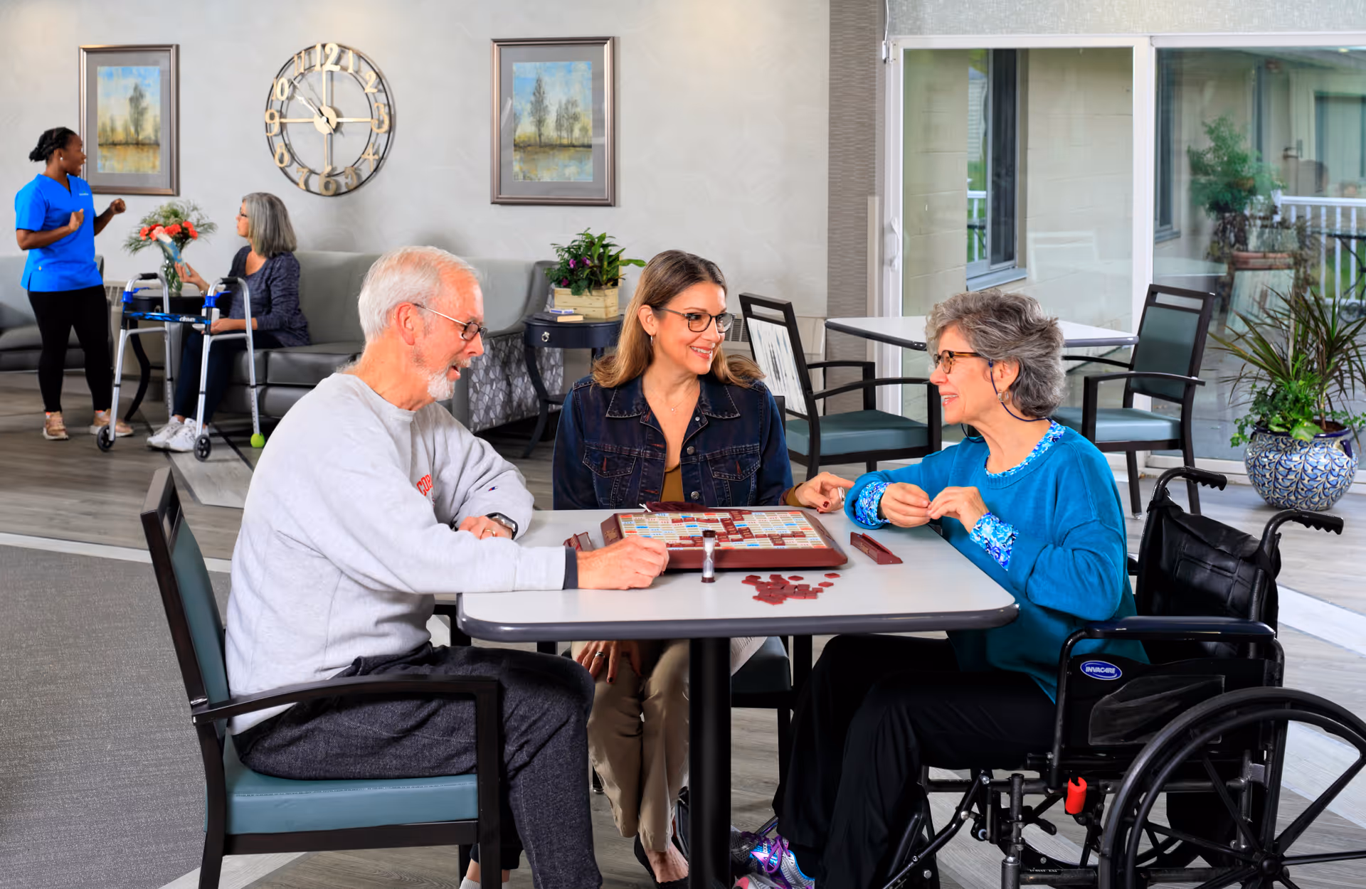 Three people, including two older adults (one in a wheelchair), sit at a table playing a board game in a bright senior living common room while staff and other residents are visible in the background.