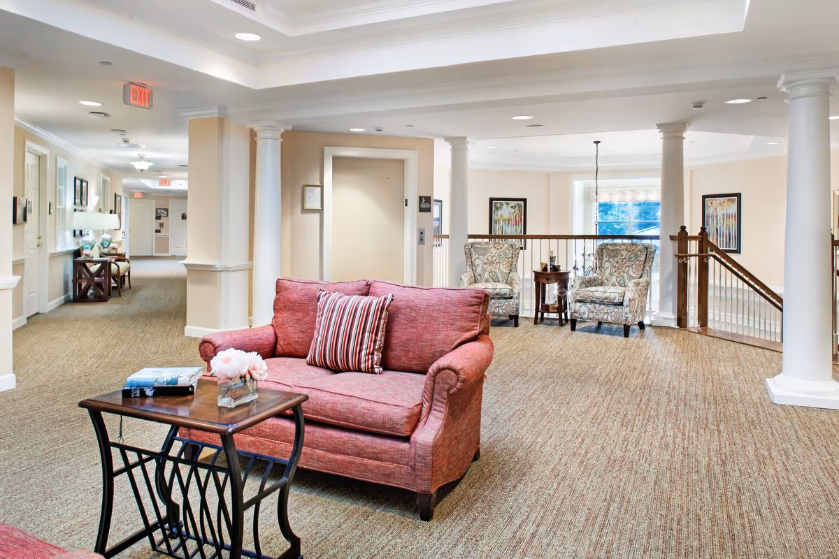 A bright and spacious common area in a senior living facility featuring a red loveseat with a striped pillow, a wooden side table with a book and a small flower arrangement, two patterned armchairs with a small round table between them, beige carpet, white columns, and framed artwork on the walls. There is an elevator and a hallway with additional seating and lamps in the background.
