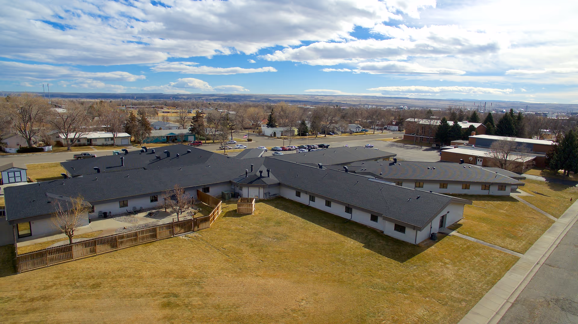 Aerial view of a single-story building complex with a dark roof and light-colored walls, surrounded by a large grassy area and a few trees. Several cars are parked in a parking lot behind the building. The sky is partly cloudy with blue patches visible.