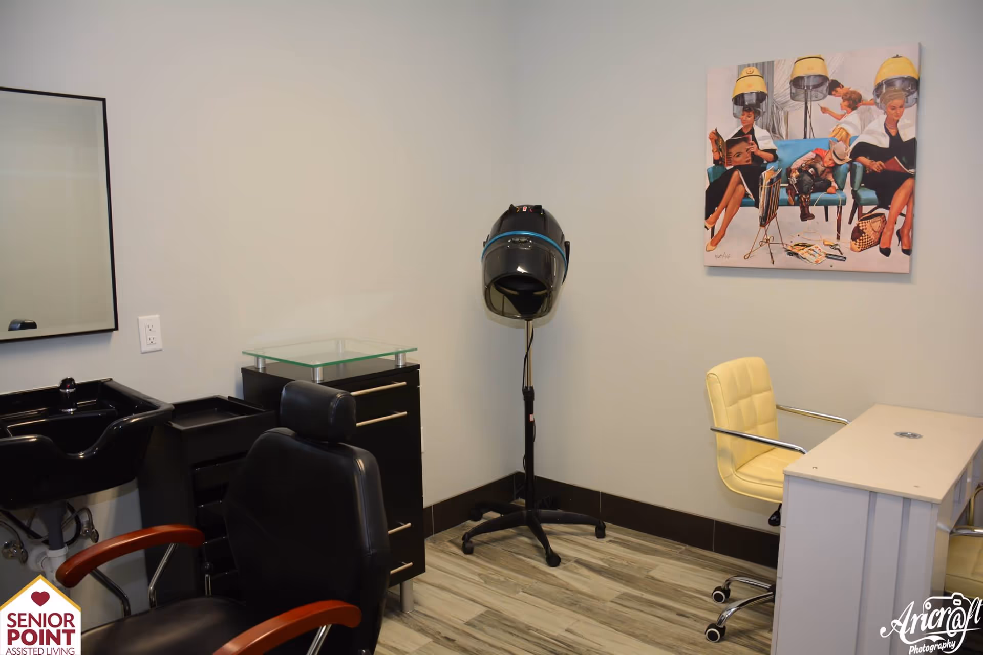 Interior of a small salon or beauty room with a black salon chair, a black sink, a black cabinet with glass top, a hair dryer on a stand, a yellow chair, a white desk, and a colorful painting on the wall depicting people sitting under hair dryers.