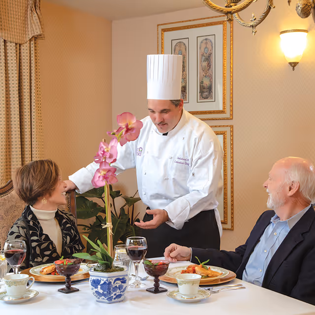 A chef in a white uniform and tall chef hat is serving two elderly people seated at a dining table. The table is set with plates of food, wine glasses, teacups, and a potted orchid centerpiece. The setting is an elegant dining room with framed artwork on the wall and warm lighting.