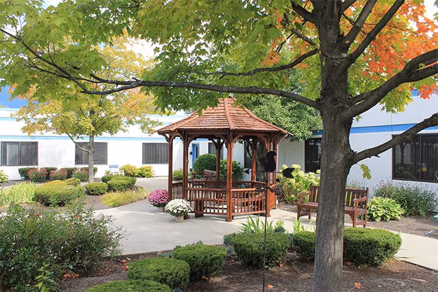 Outdoor garden area at Elderwood Assisted Living at Tonawanda featuring a wooden gazebo surrounded by green shrubs, trees with some autumn-colored leaves, and benches along a paved walkway in front of a white building with blue trim.