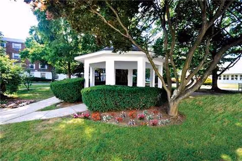 A white gazebo sits among manicured shrubs and a tree-lined lawn outside the facility.