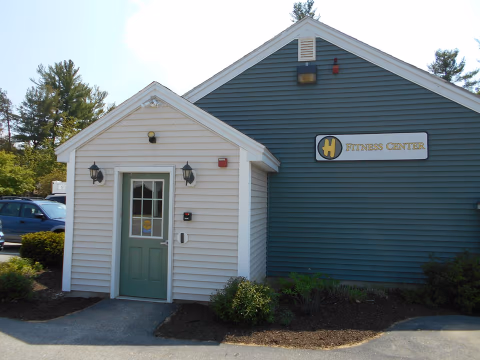Exterior view of a building with white and blue siding, featuring a green door with glass panels. There are two black wall-mounted lanterns on either side of the door. A sign on the blue siding reads 'Fitness Center'. Surrounding the building are some bushes and trees, with a parked blue vehicle visible in the background.