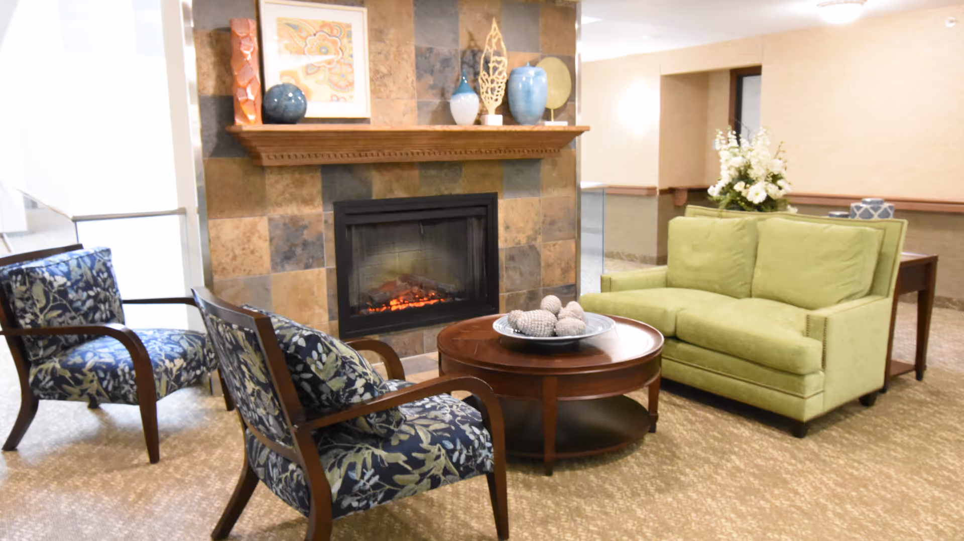 Cozy seating area with a fireplace, a green loveseat, two patterned armchairs, and a round coffee table with decorative objects.